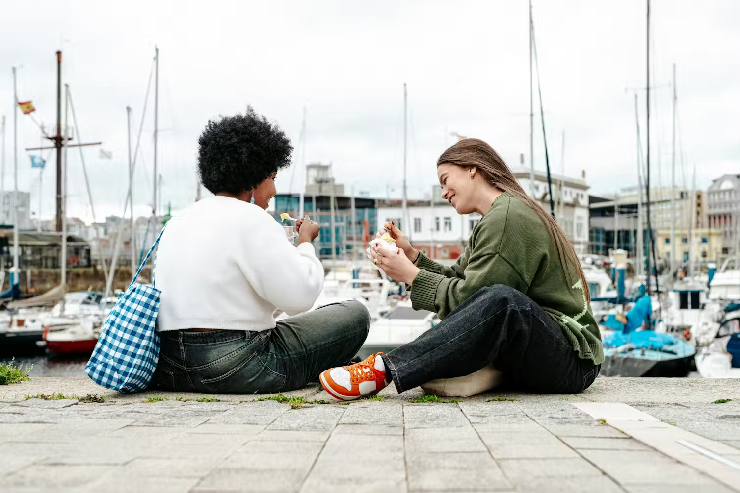 Two women sit on concrete, with a harbour and boats behind them.