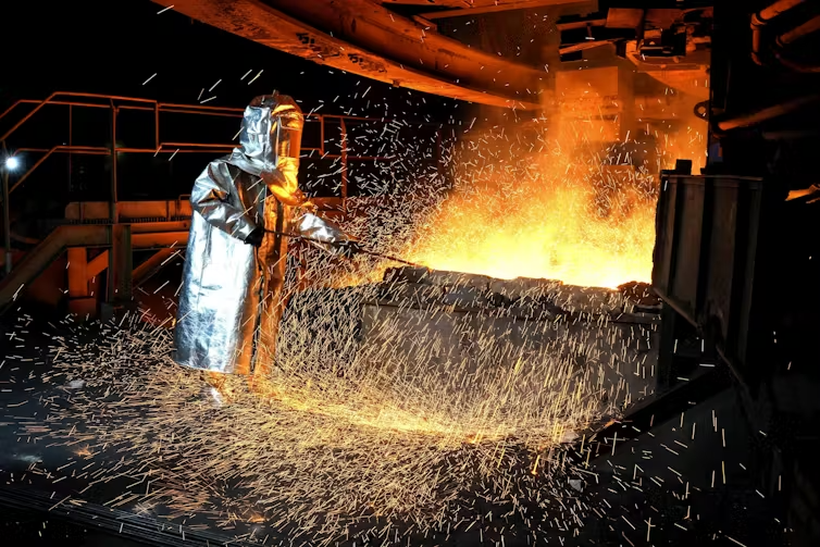 A worker in a protective suit pokes a metal rod to tap slag from a smelting furnace.