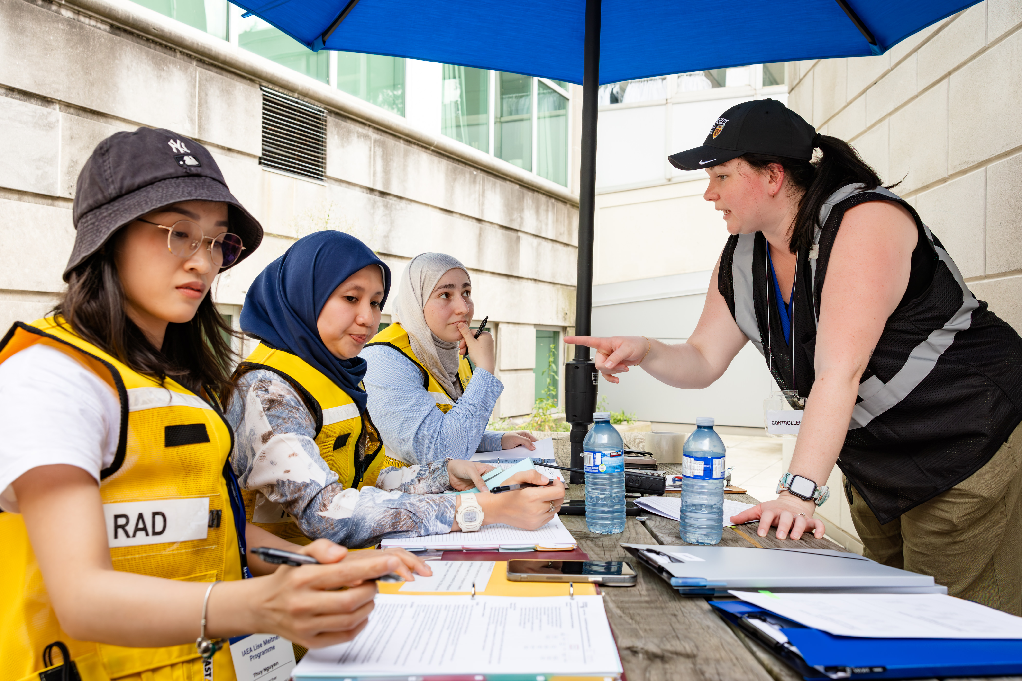 Three women sit at a picnic table, while another women leans over the picnic table and gives them instructions.