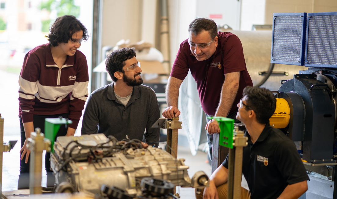 Professor Ali Emadi leans over a table, chatting with 3 graduate students.