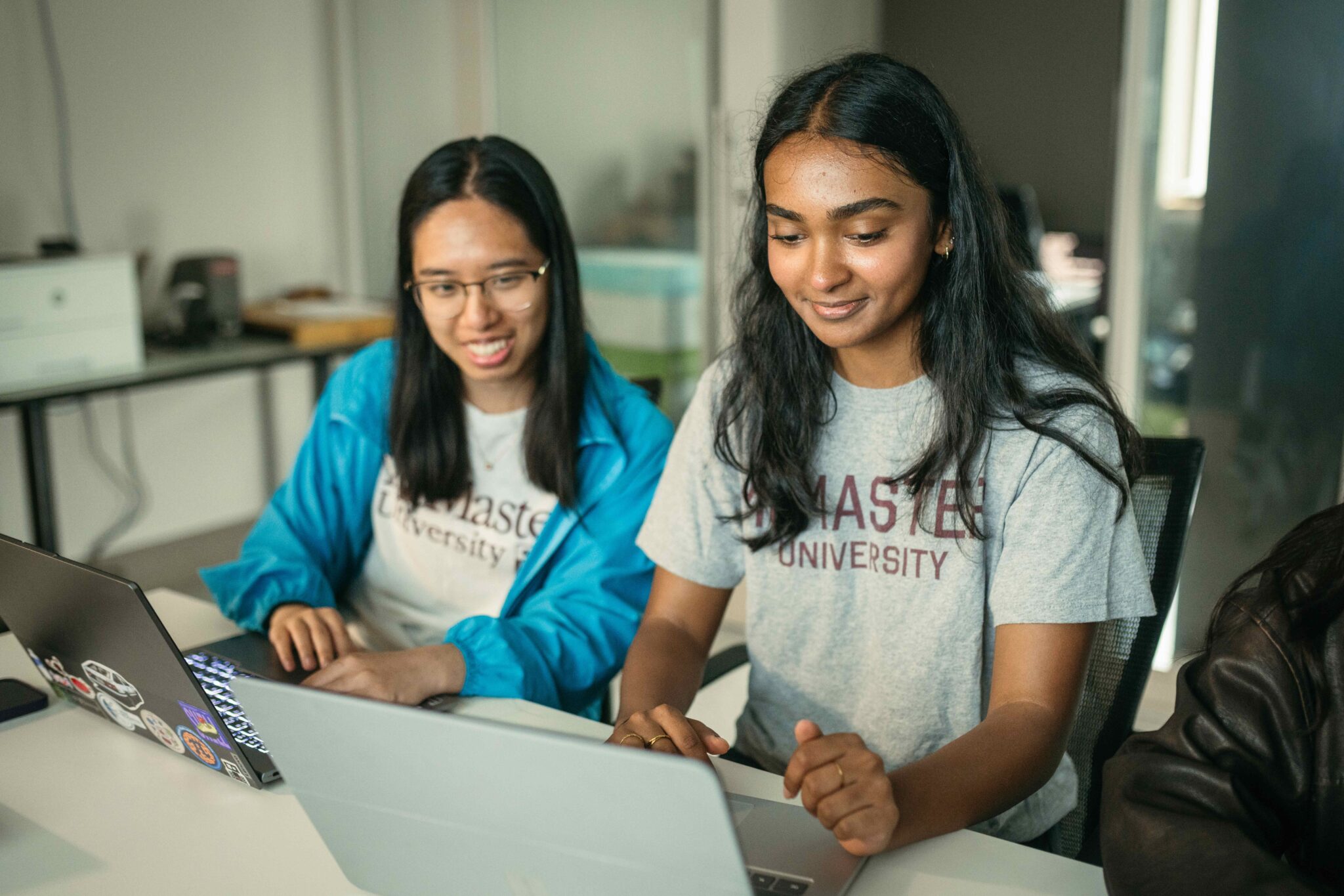 Two young women sit ata table, looking at laptops open on the desk in front of them.
