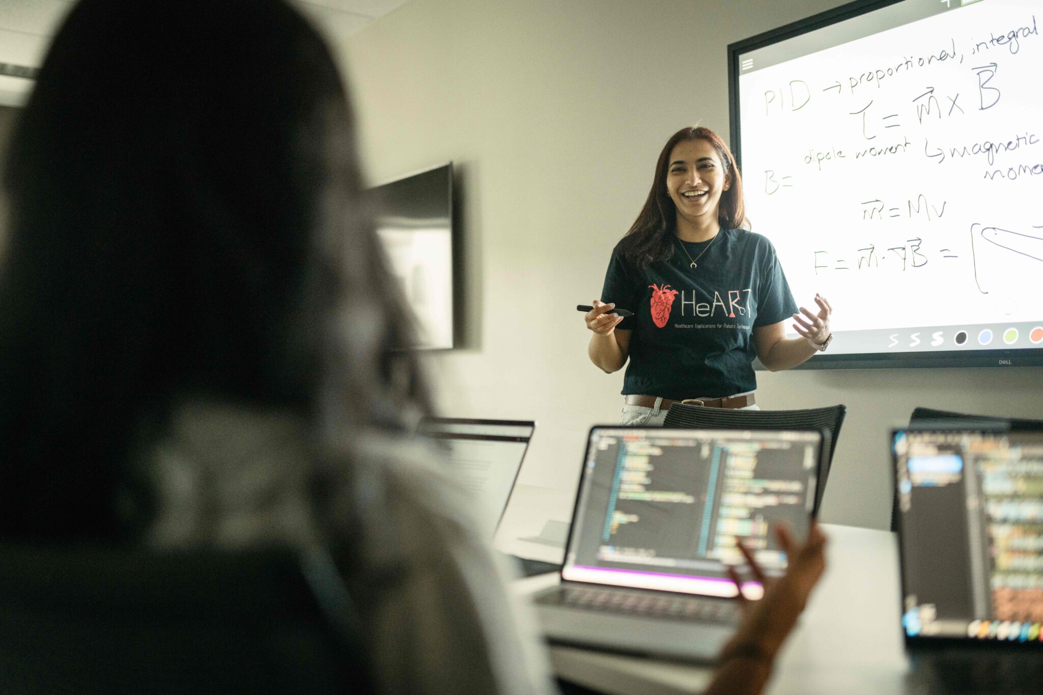 Looking over the shoulder of somebody sitting at a desk, with their laptop open, and several other laptops beside them, there is a woman standing at the front of the room, holding a marker and gesturing. On a large whiteboard behind her are several words and equations. 