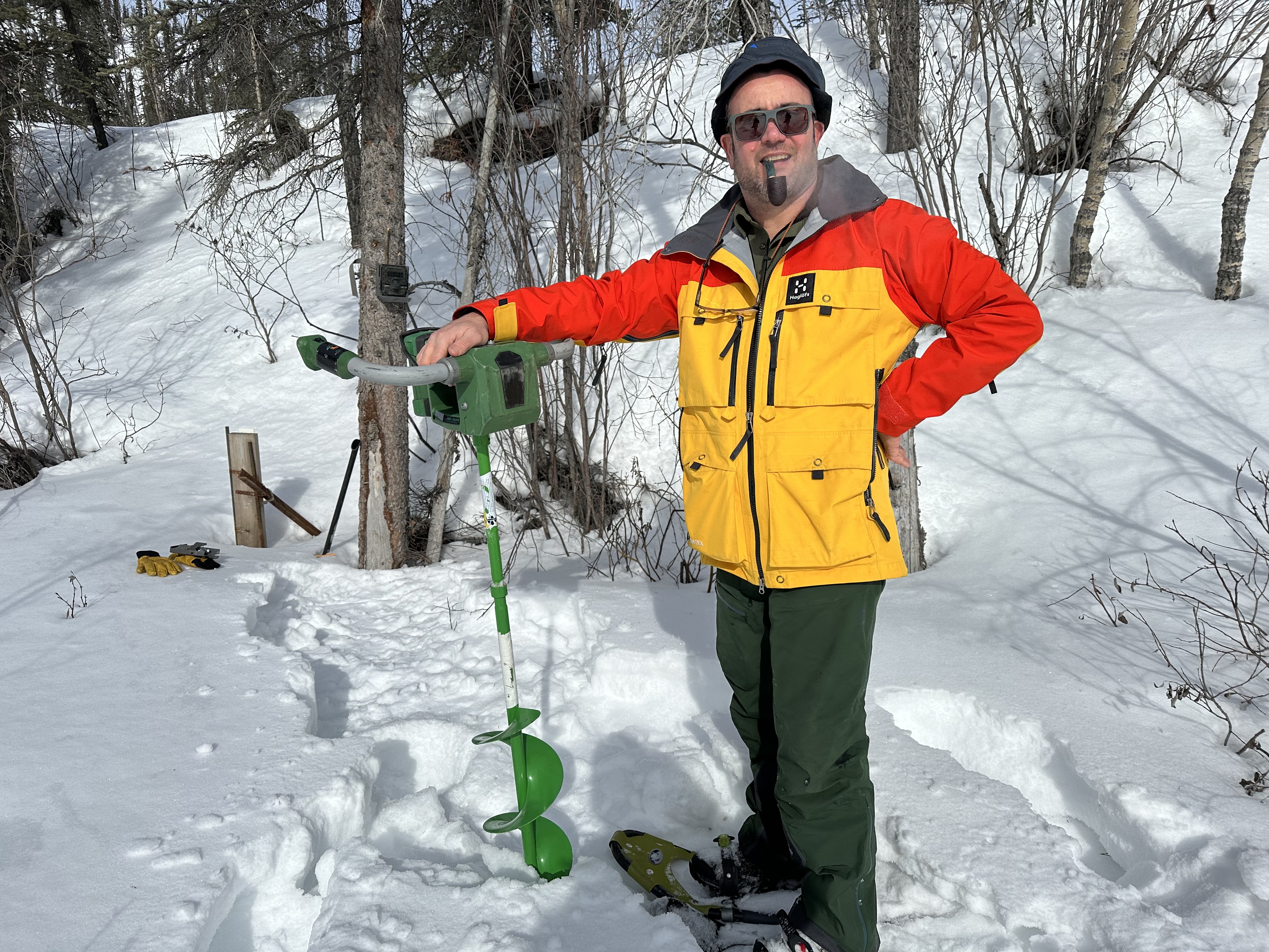 A man in a winter coat stands in the snow, holding the end of a large green drill that's going into the earth. He is holding a pipe in his mouth.