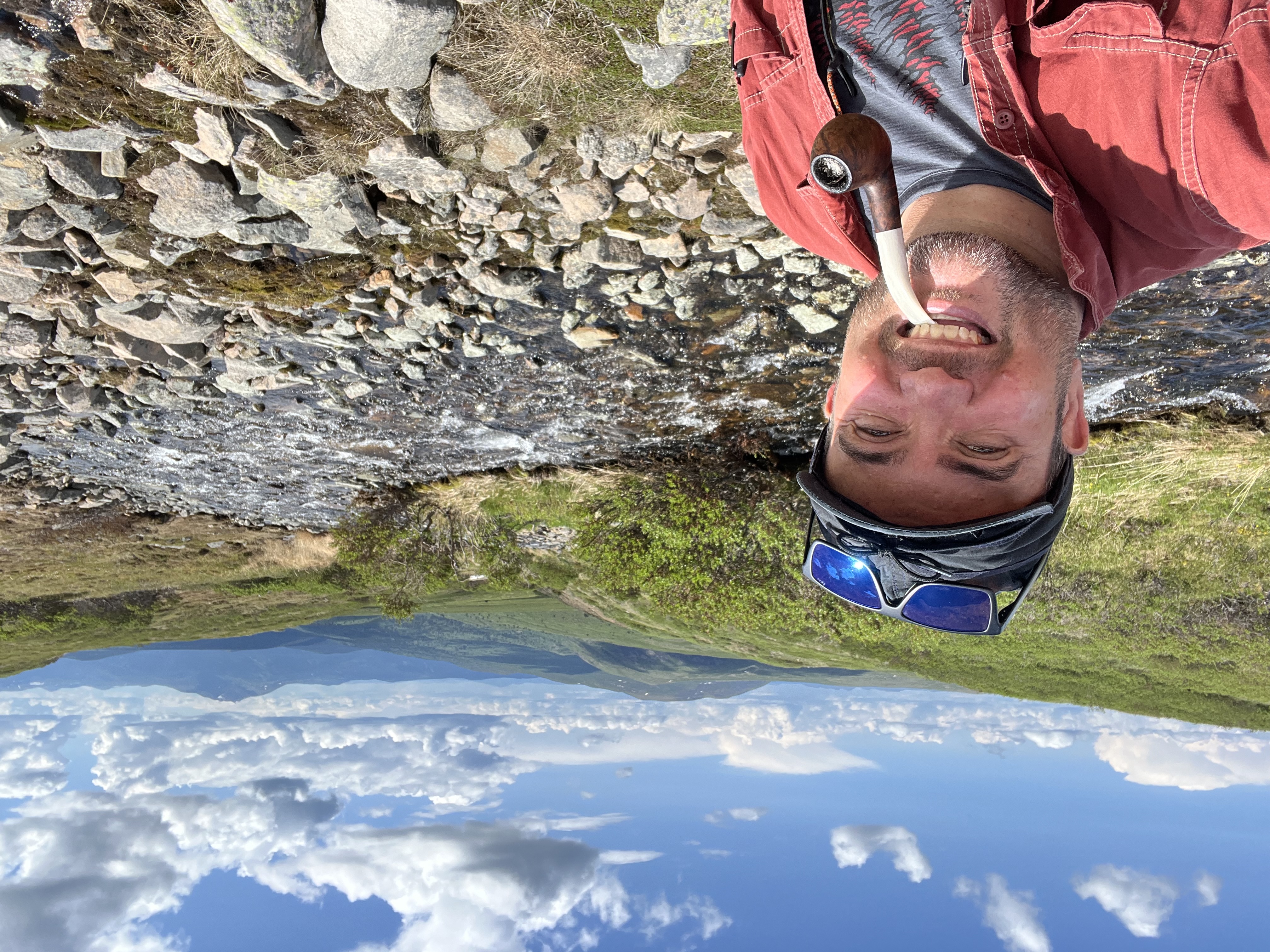A man holds a pipe in his mouth, taking a selfie in front of a stream. Rolling hills and mountains are behind him.