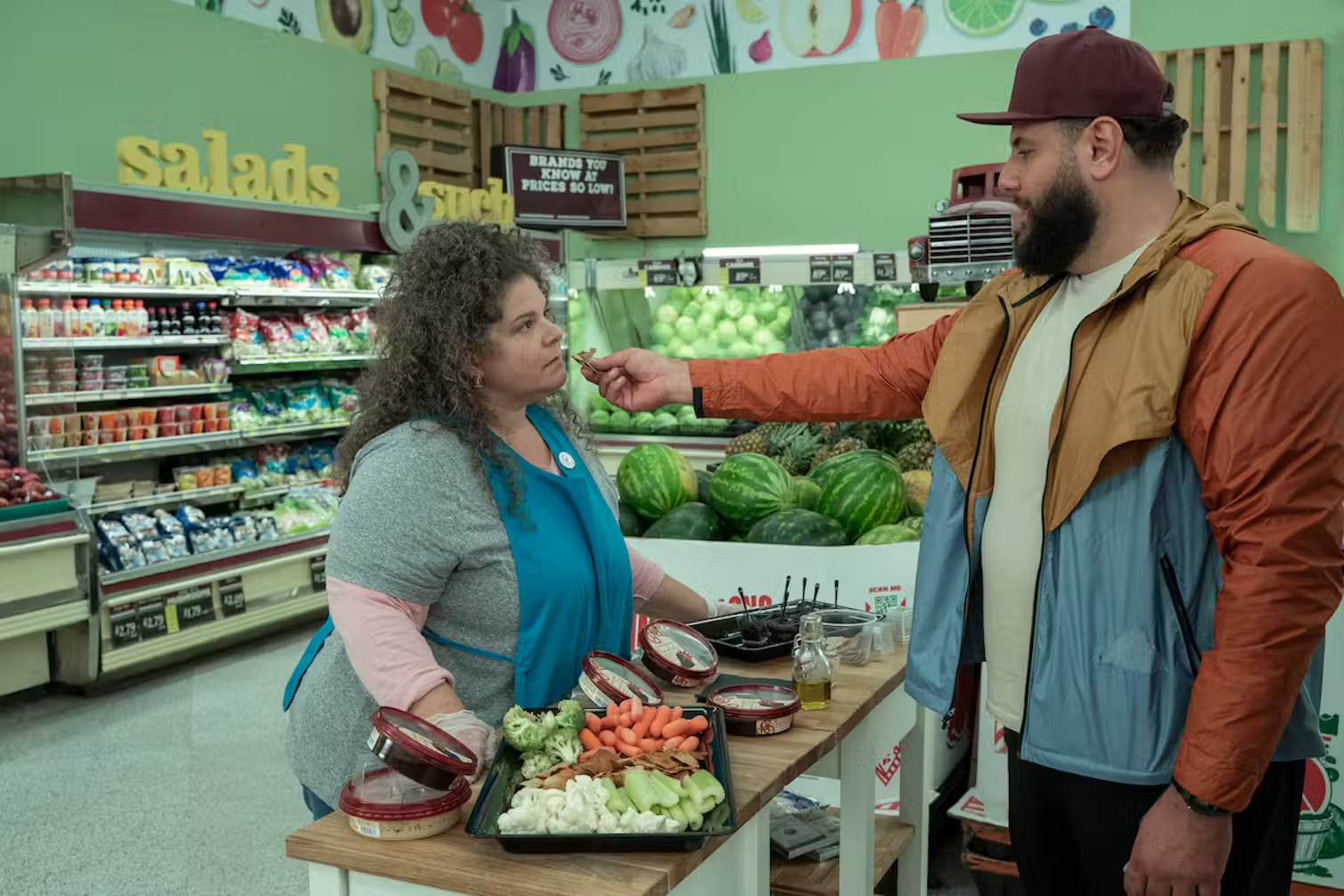 In a grocery store, a man holds out an olive to a woman working.