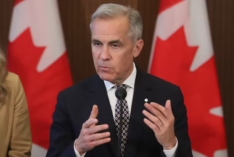 Prime Minister Mark Carney speaks at a press conference, in front of two Canadian flags.