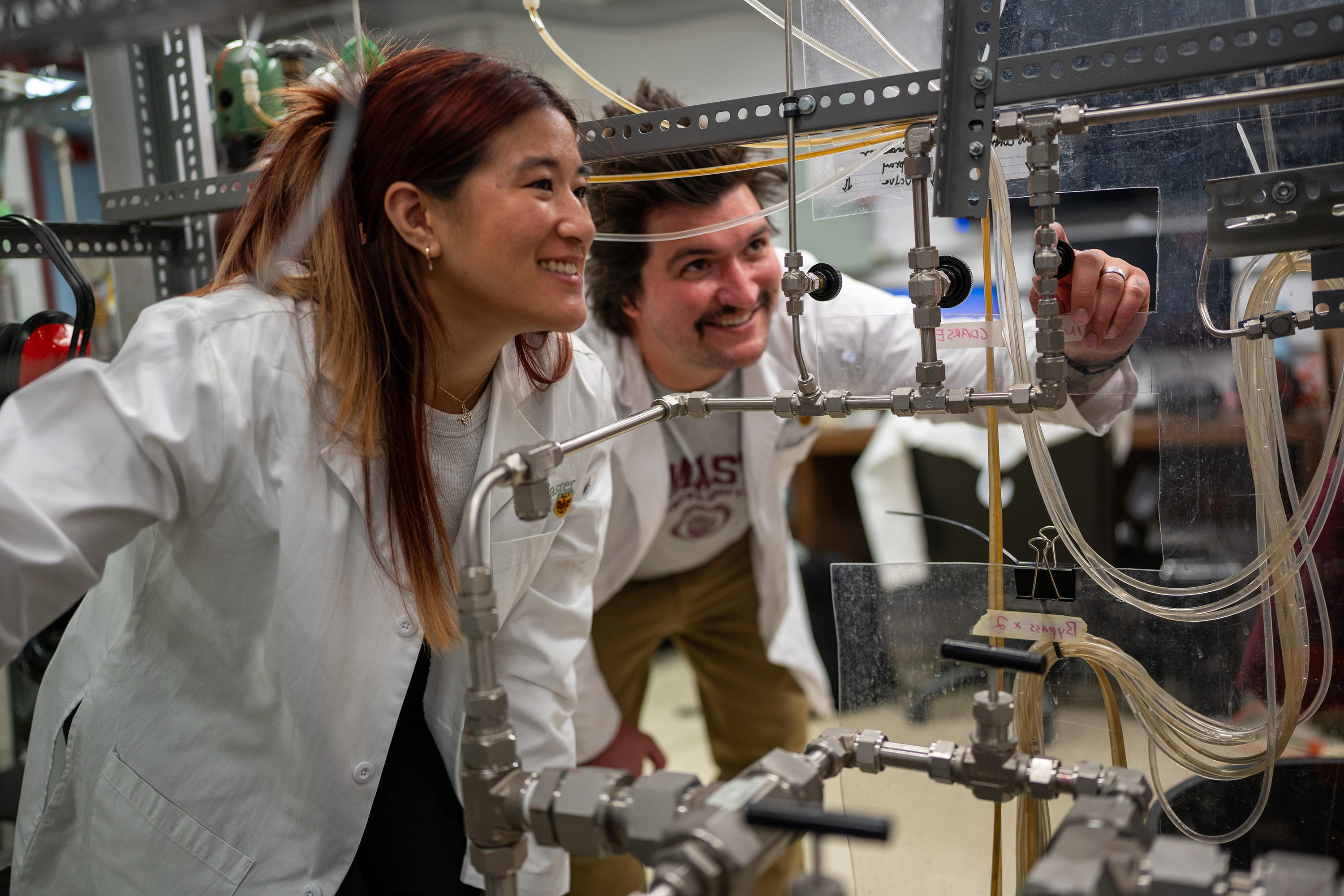 Two people in lab coats look at various taps and coils in a lab.