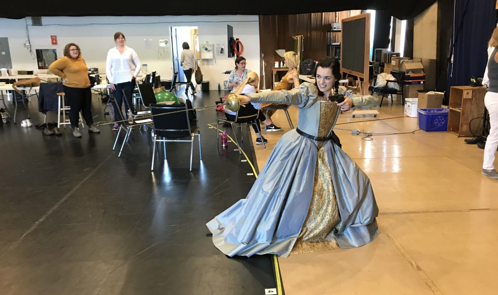 An actor in a blue dress lunges with a sword