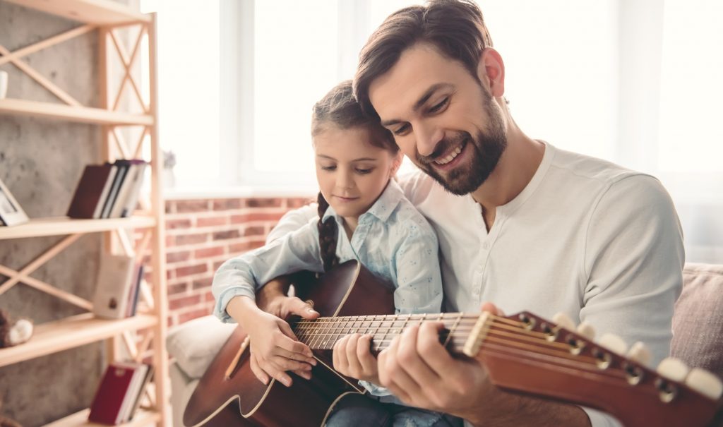 A smiling man is teaching a smiling little girl on his lap to play the guitar.