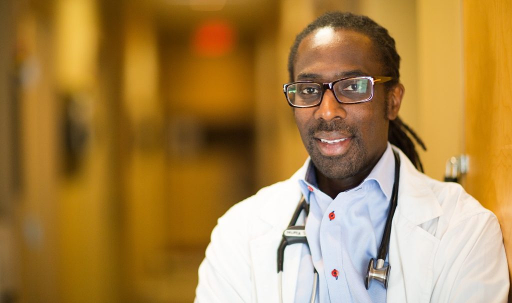 A head-and-shoulders image of Sean Wharton, looking into the camera, wearing a lab coat and has a stethescope around his neck
