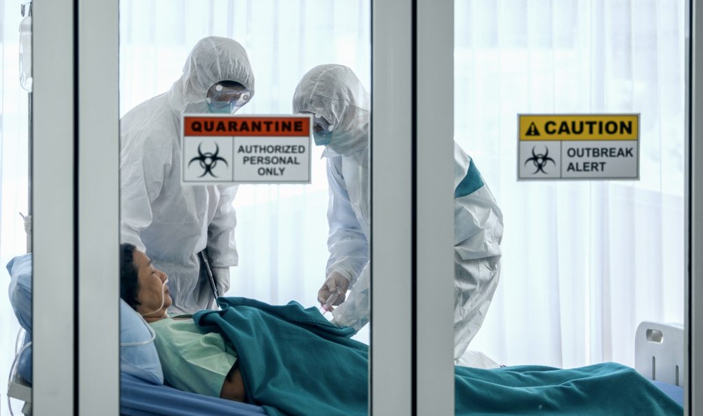 A patient lies in a hospital bed behind glass doors that are closed and labelled with quarantine signs. Two medical personnel in full-body protective suits are tending to the patient.