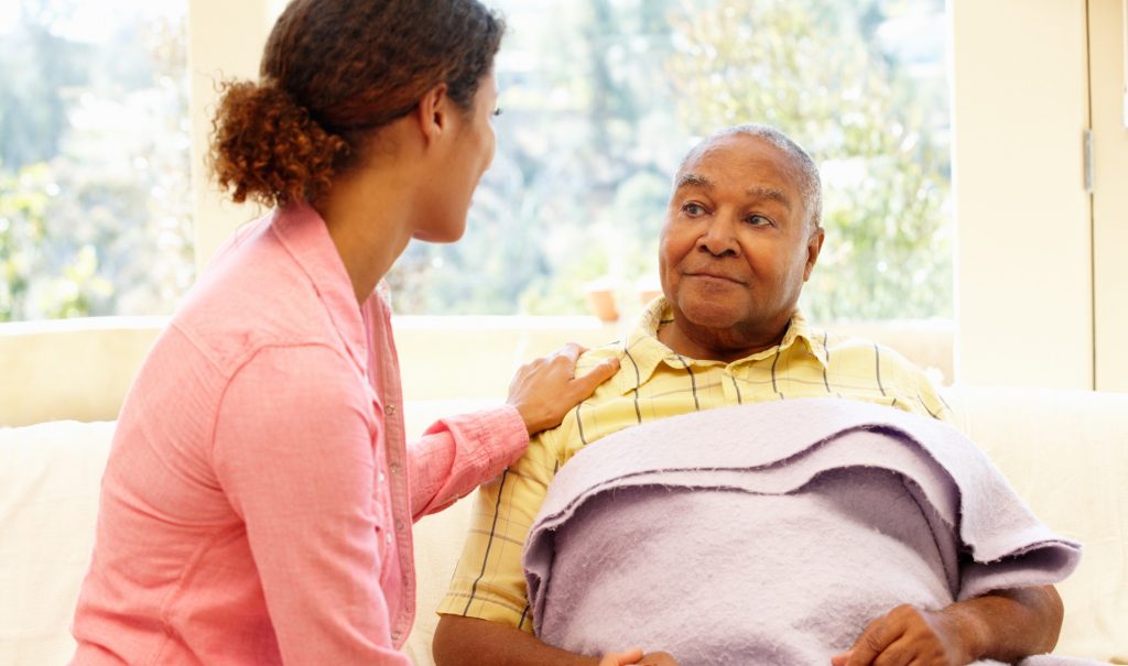 A woman sitting beside an older man sitting up in bed with a blanket. She has her hand on his shoulder and they're both half-smiling and comfortable looking