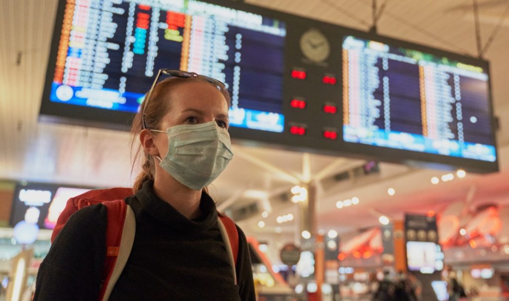A woman wearing a face mask in the foreground, with an airport arrival/departure chart above her in the background.