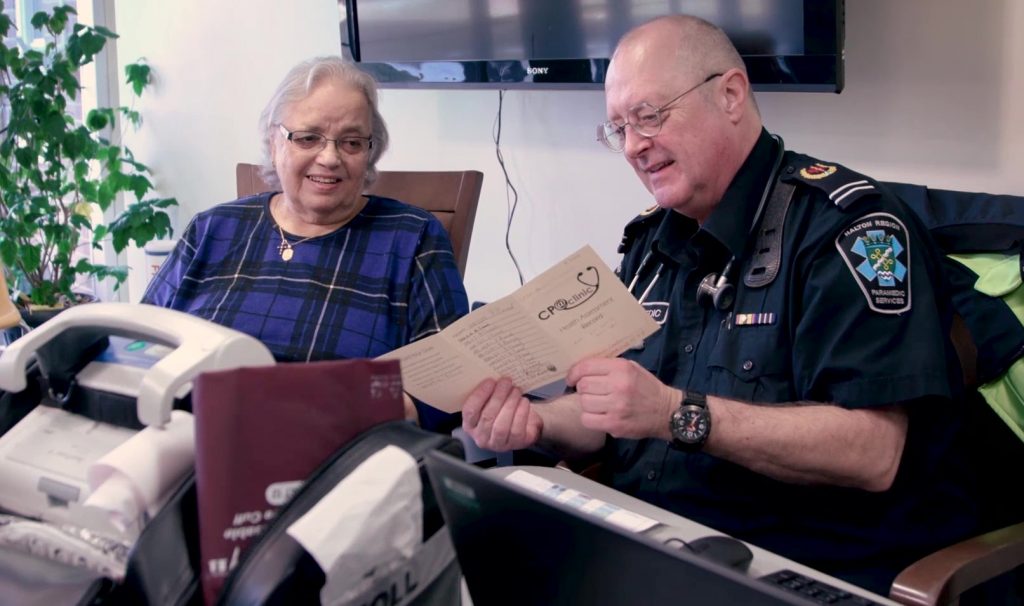 An older woman and a man as a paramedic are sitting together in a home setting. It looks like the paramedic is reading to the woman. Both are smiling.
