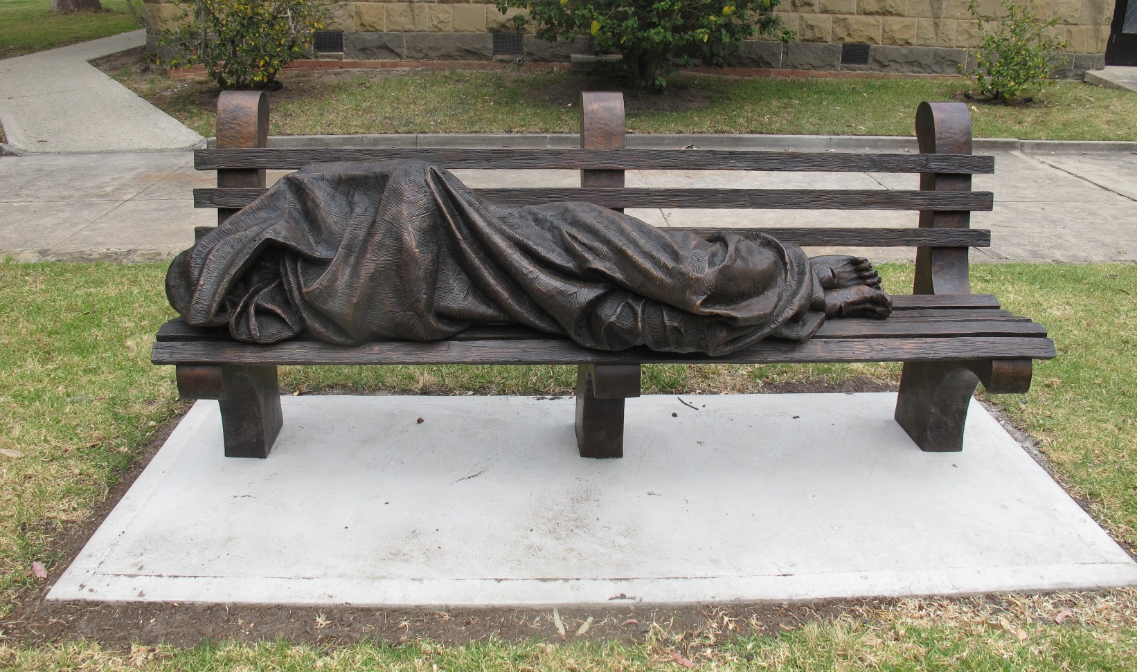 A dark bronze, realistic-looking statue of a person wrapped in a too-small blanket lying on a public bench.