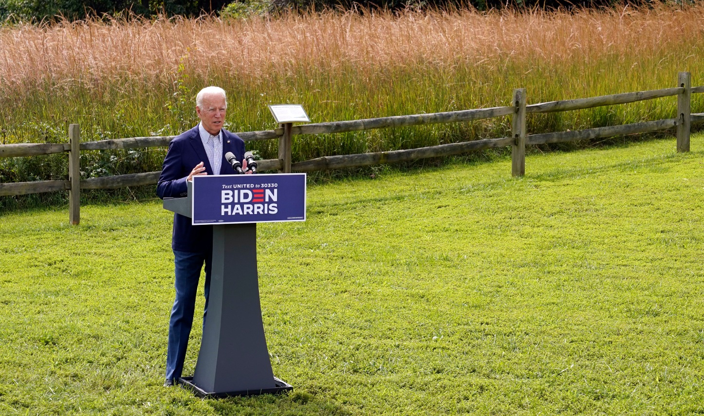 Joe Biden giving a speech from a podium in a grassy field in the sunshine.
