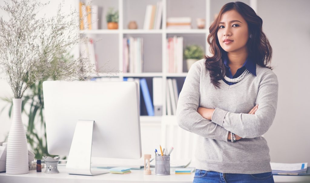 A young woman is standing with her arms crossed, confidently, in front of a desk. There's a computer and office supplies on the desk, and a large, well-organized shelf full of files and books in the background.