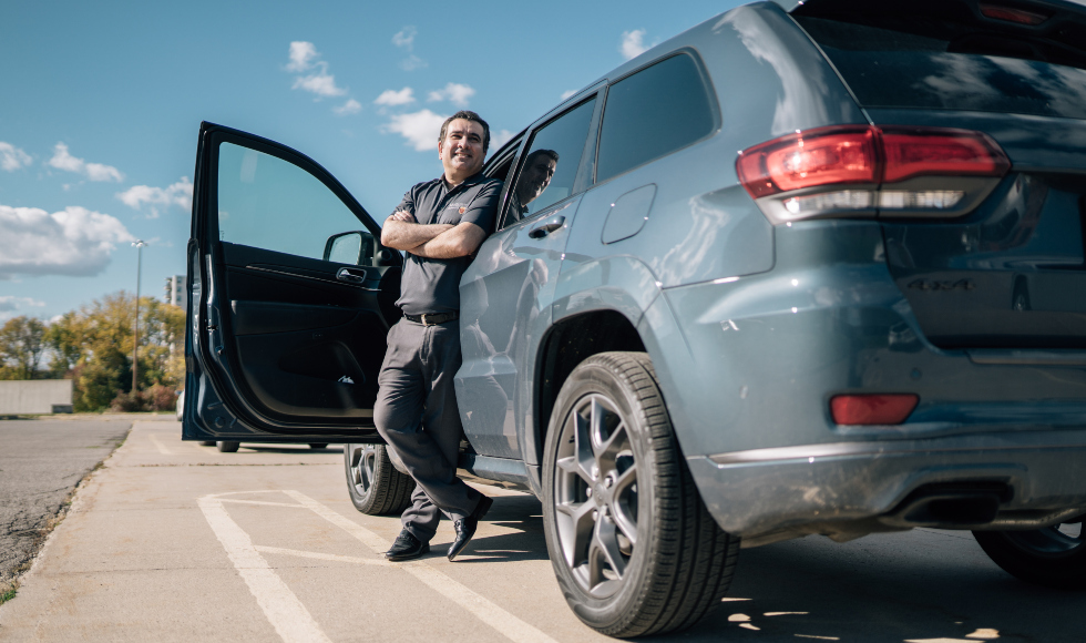 A smiling man stands outside in front of a minivan with its door open. The sky is blue and the sun is shining.