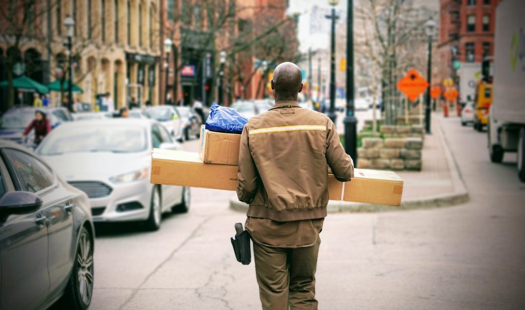 A delivery person or shown from behind carrying cardboard boxes down the sidewalk.