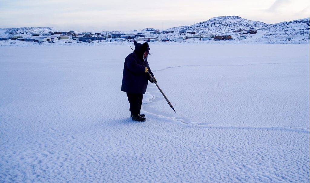 A figure in a parka stands on snow-covered sea ice in the foreground, testing it with a pointy object. Far on the other side of the ice in the background, a lot of low, snow-covered buildings.