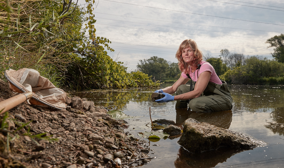 A woman wearing hip-waders crouches in shallow water, holding a clump of dirt