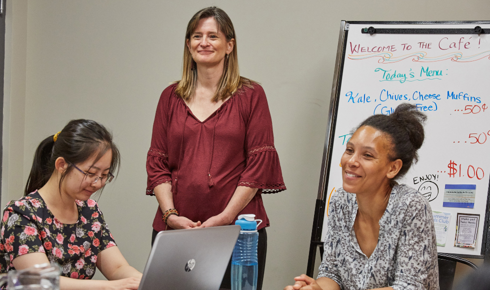 A woman in a red shirt stands smiling between two other seated smiling women. There is a whiteboard in the background