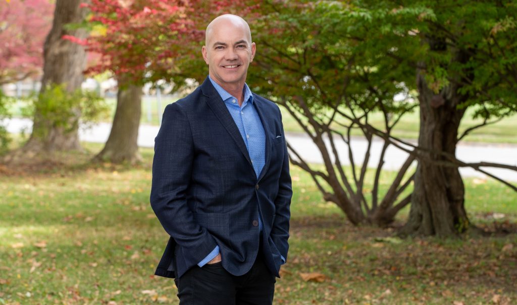 Marty Gibala wearing a suit smiling into the camera with his hands in his pockets against a backdrop of trees in the fall.