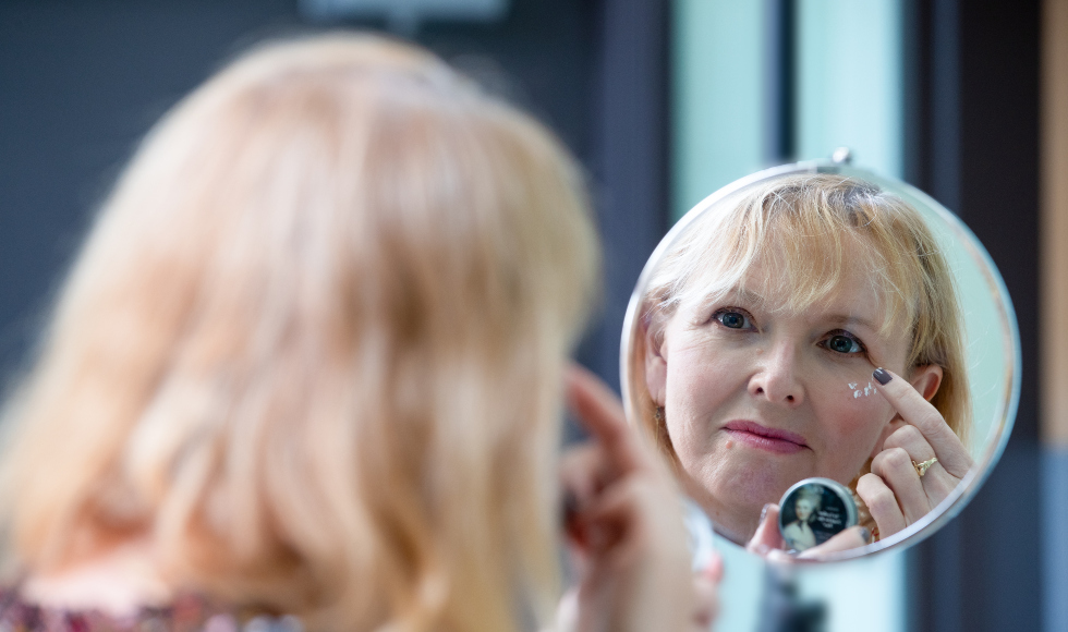 A blond woman is reflected in a mirror. She is putting white cream on under her eyes.