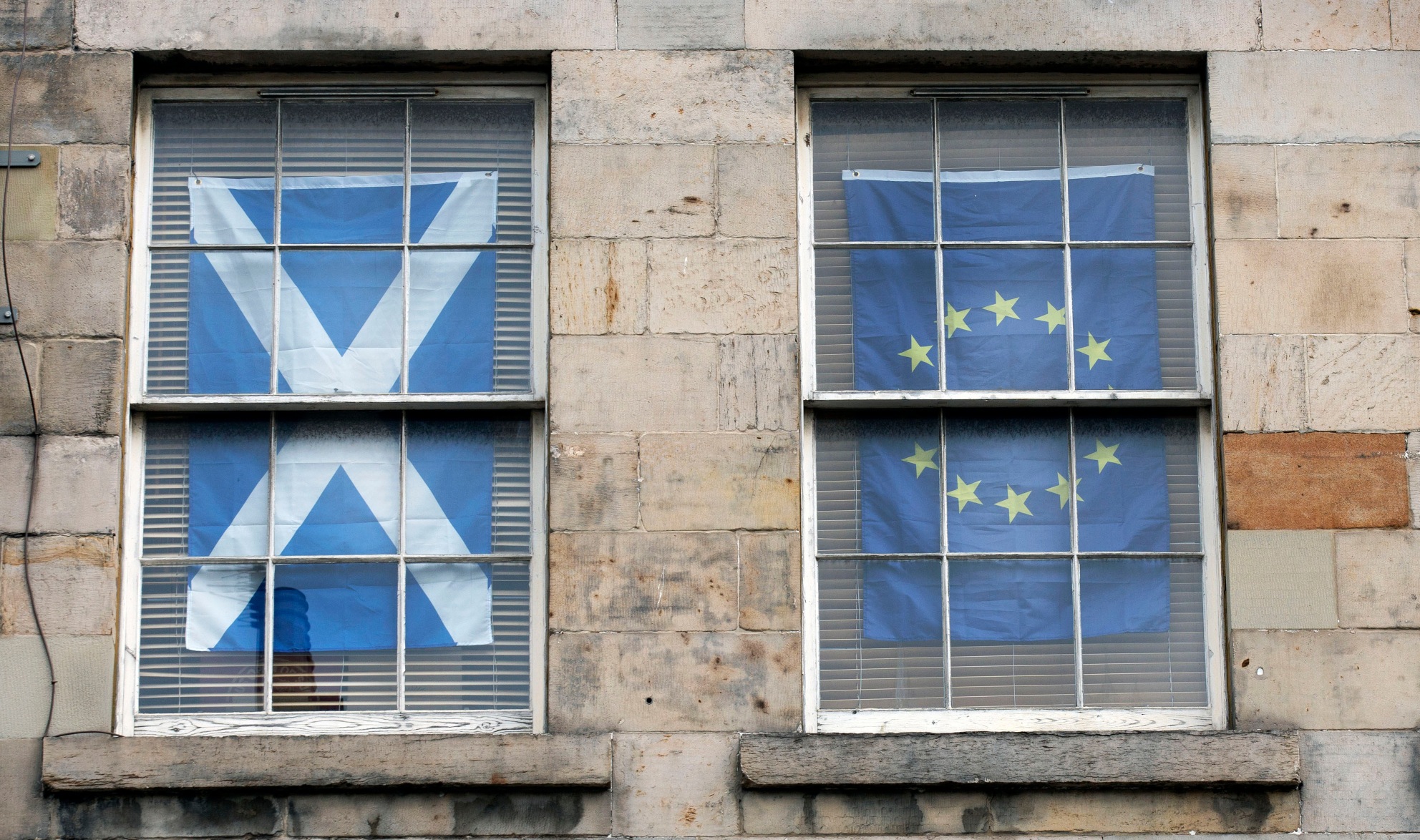 Two apartment windows, side by side: one is curtained by a Scottish Saltire flag, the other by an EU flag.