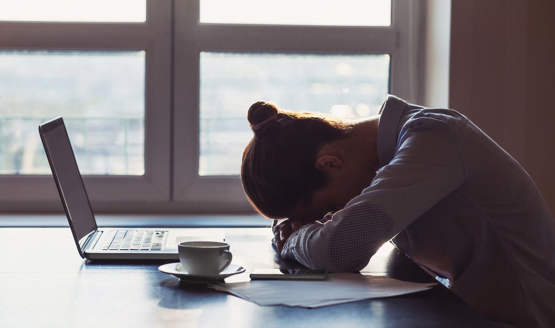 Silhouette of a person sitting in front of a laptop — they seem discouraged, with their head resting on their arms on the desk.