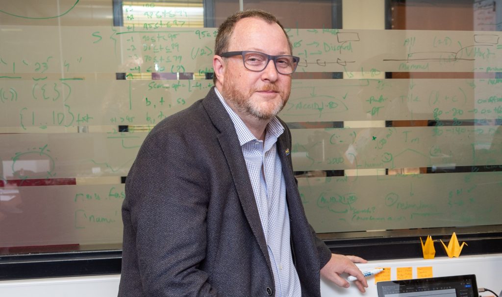 Andrew McArthur looking into the camera, holding a marker with a glass surface behind him covered in writing.