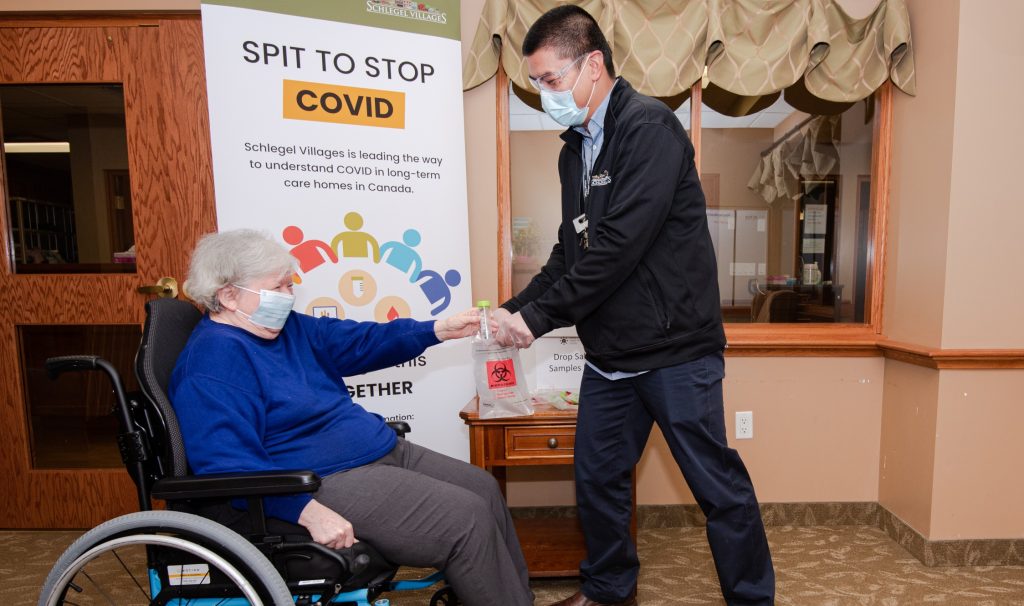 An older woman wearing a mask in a wheelchair hands a vial to a medical worker. They're in front of a sign about spit tests for COVID