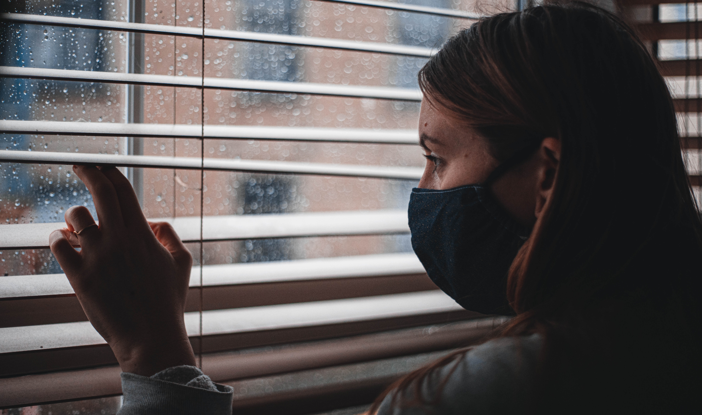 A person wearing a mask looking out between window blinds