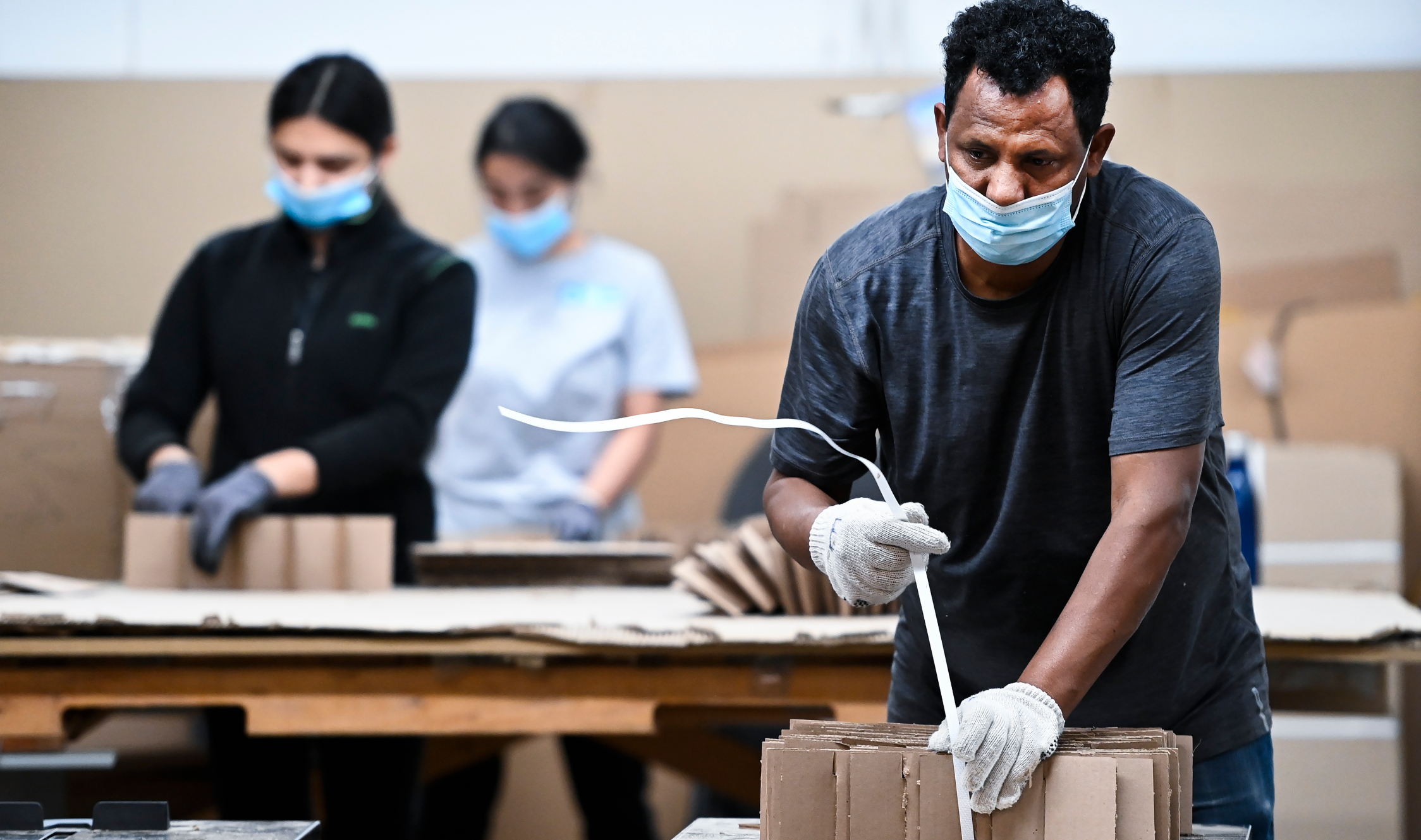 Masked, gloved workers package cardboard objects.