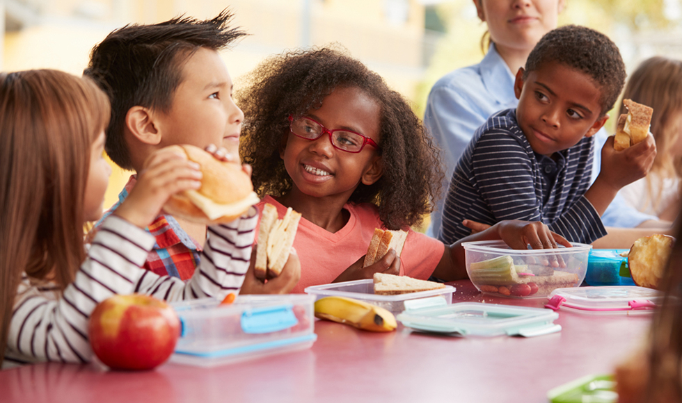 A group of schoolchildren eating lunches out of lunchboxes at a table.