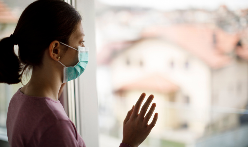 Woman wearing a mask, looking out a window with her hand raised as though to touch the window or wave.