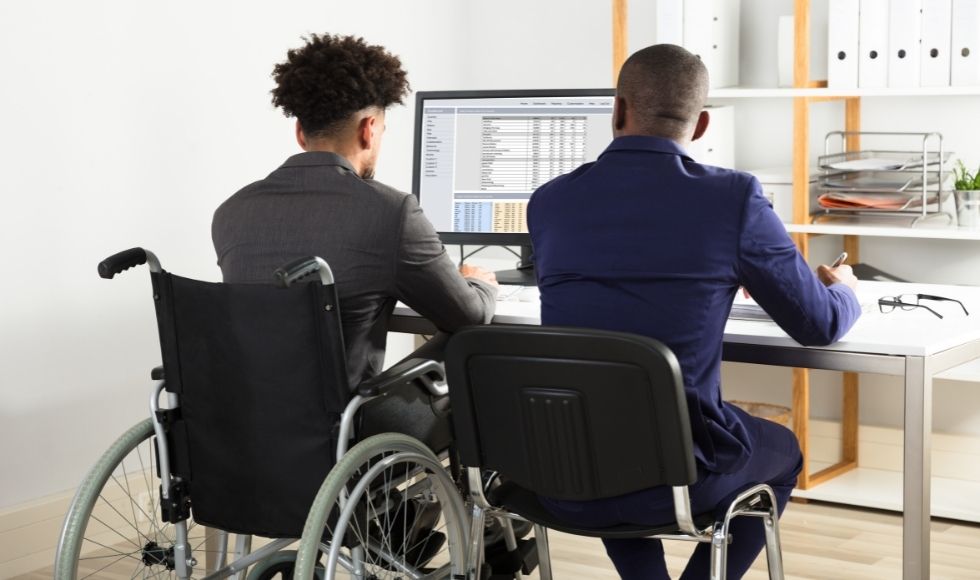 Two people seen from behind, sitting at a desk with a computer. One is in a wheelchair.
