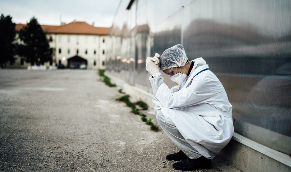 A health-care worker wearing personal protective equipment is bent over with their head resting on their hands.