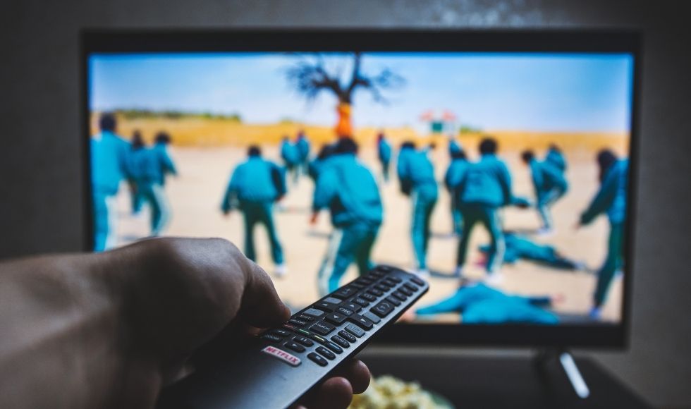 Hand holding remote in foreground in front of a big screen showing Squid Games miniseries