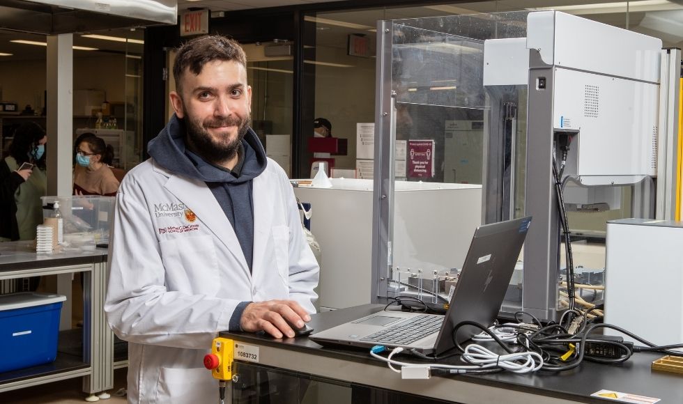 A man in a labcoat standing at a desk with a computer on it in a lab setting. He's smiling at the camera,