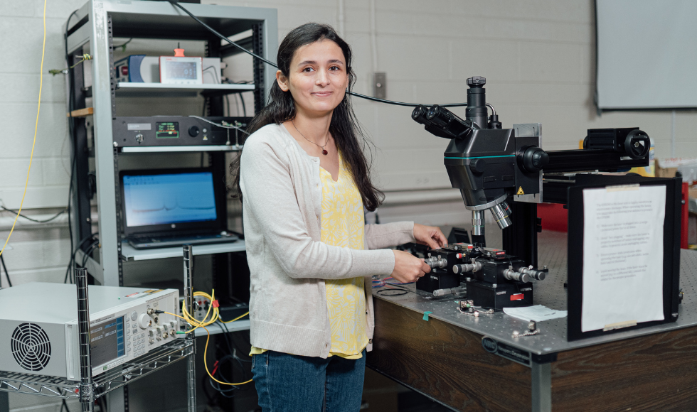 Engineering Physics PhD student Khadijeh Miarabbas Kiani smiles at the camera. She is standing inside a laboratory and has equipment all around her.