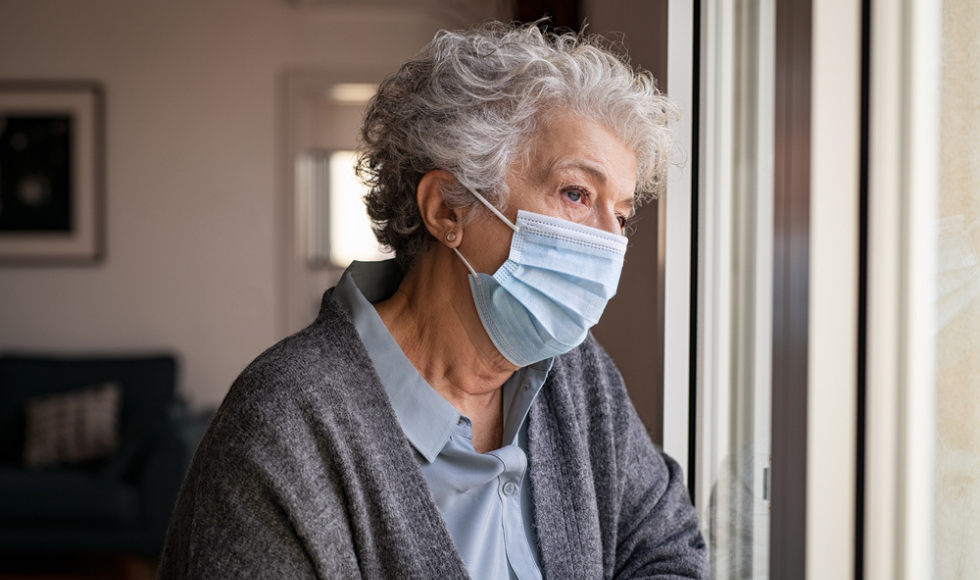 An older woman with grey hair, wearing a grey sweater and blue collar top looks out a window. She is wearing a mask.