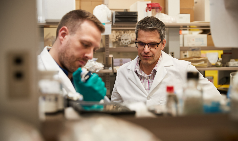 A picture of two men wearing lab coats. One of the men is injecting something that is blocked from view, while the other man watches. The man watching is Brian Coombes, a McMaster biochemistry and biomedical sciences professor.