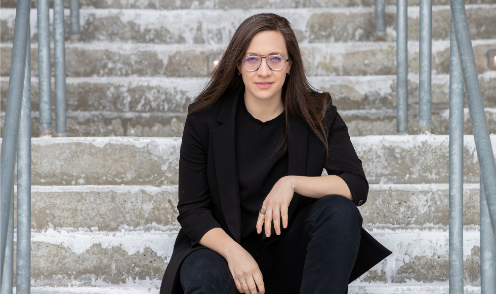 A picture of Vass Bednar sitting on a stone staircase. She has her arms resting on her thighs and is staring straight into the camera.