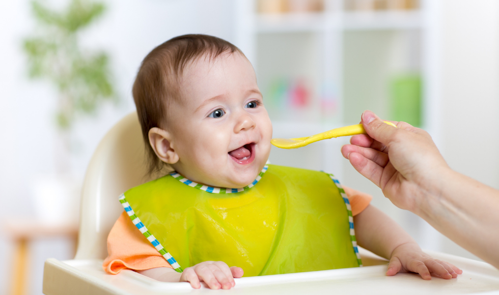 A picture of a smiling baby wearing a green bib and sitting in a highchair. An arm holding a yellow plastic spoon is in the lower right corner of the image.