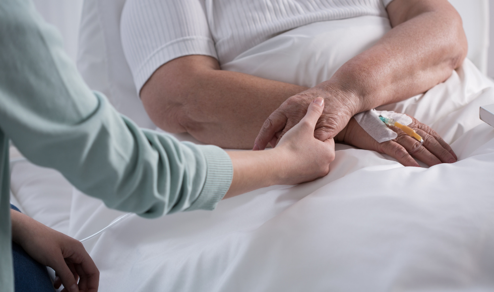 A photo of the arms of a person in a bed. Another person's arm is reaching out to them, holding one of their hands. One of the hands of the person lying in bed has an IV needle in it. No faces are in the frame.