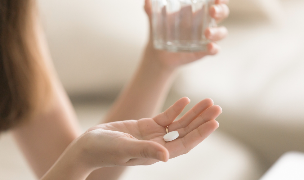 A photo of a woman’s hands. She is holding a glass of water in one, and a white pill in the other. Some of her hair is in the top left corner of the shot, but her face is not visible.