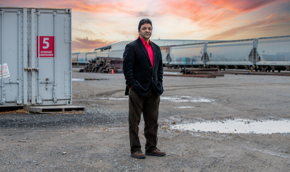 A photo of Manish Verma standing in a rail yard. There is a train in the background over his left shoulder, and a shipping container over his right shoulder. He is wearing a pink dress shirt, a black suit jacket and brown pants and shoes. He is looking directly at the camera and not smiling.