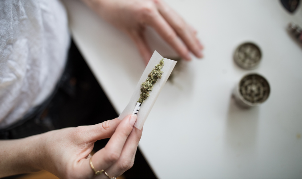 A picture of a woman’s right hand holding a marijuana cigarette that has not yet been fully rolled. Her left hand is out of focus and resting on a table top in the background. Her face is not visible. There are some cannabis-related items on the table.