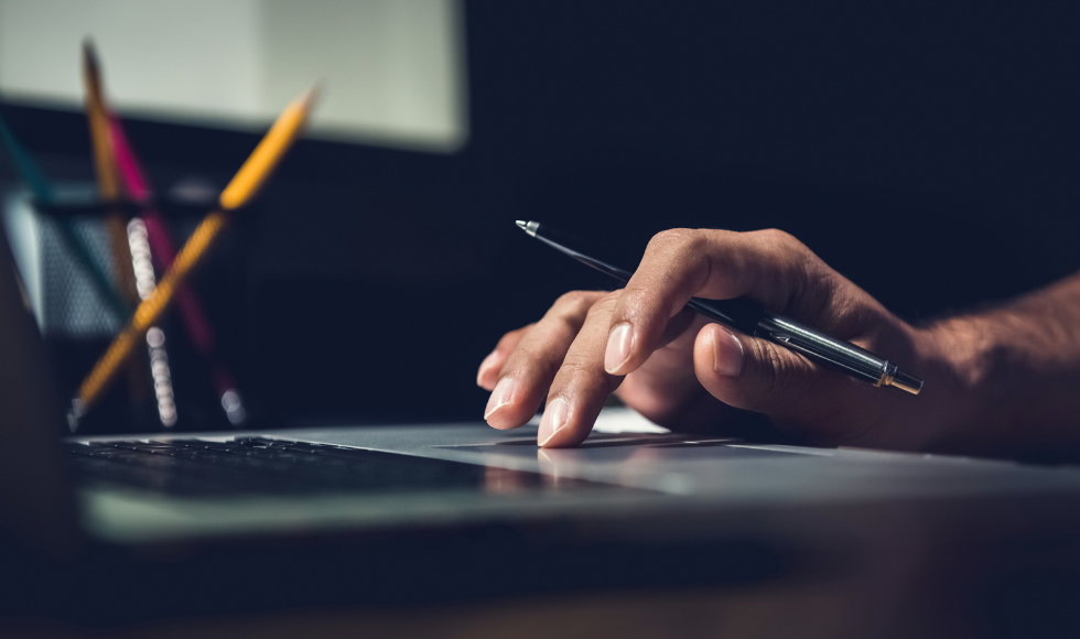 A photo of a person's hand holding a pen while resting on a laptop. The background is mostly dark, but a container of pens and a notebook can be seen beside the laptop.